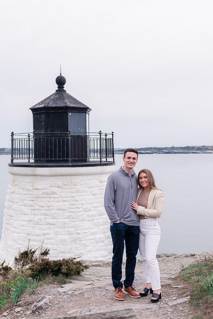 couple stands near lighthouse after castle hill surprise engagement